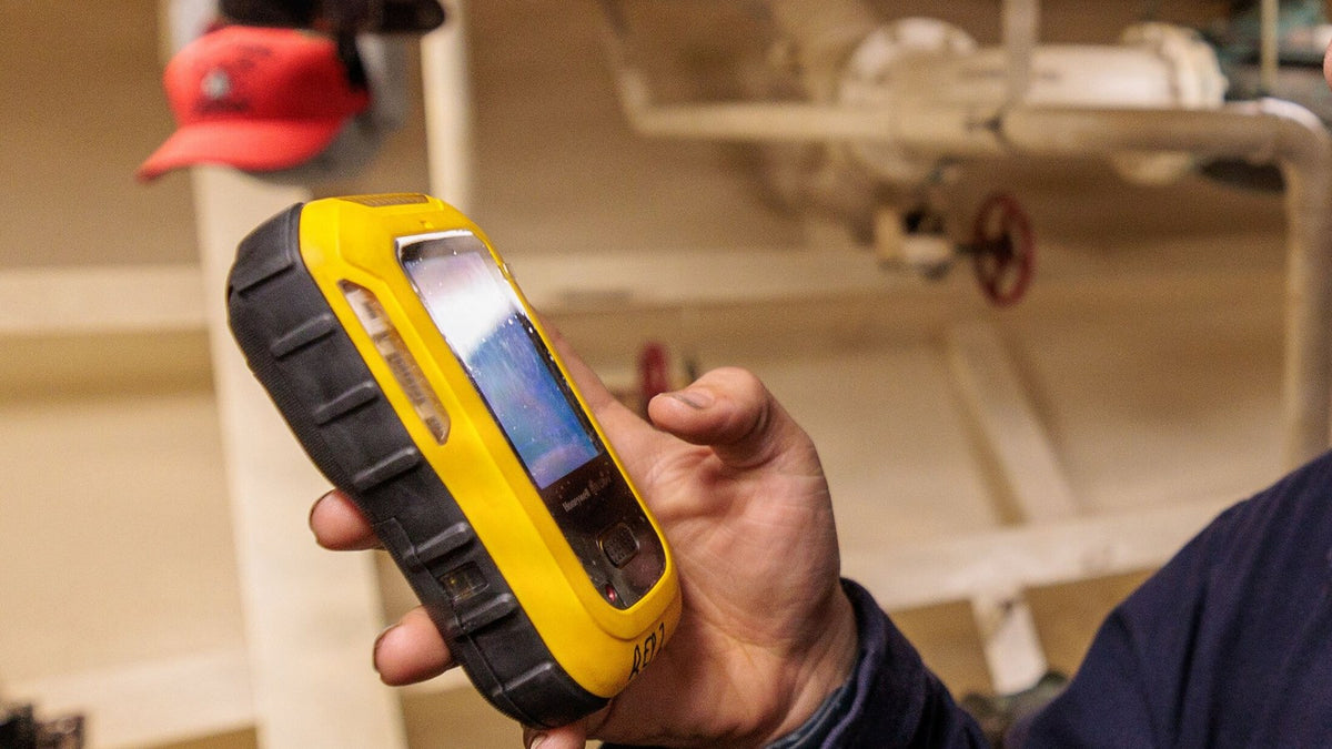 Hand holding a portable 4-gas detector in a plant room with pipework